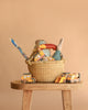 The Easter Basket Gift Set sits on a wooden stool, filled with toy vehicles, a plush bear, a Maileg Rabbit, and a long candy stick. Two Construction Toy Cars are placed in front of the basket. Neutral beige background.