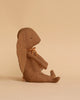 A Maileg Bunny - Chocolate Brown with long floppy ears is sitting upright against a matching beige background. The soft bunny has a simple design with stitched details for the eyes and nose, and a small bow tied around its neck. Crafted from recycled polyester, it even has magnets in its hands for playful posing.