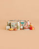 A Konges Sløjd Mini Market Grocery Set featuring a miniature shopping basket surrounded by tiny toy groceries—cereal boxes, milk bottles, eggs, pastries, waffles, a cake, and juice—all displayed against a plain beige background.
