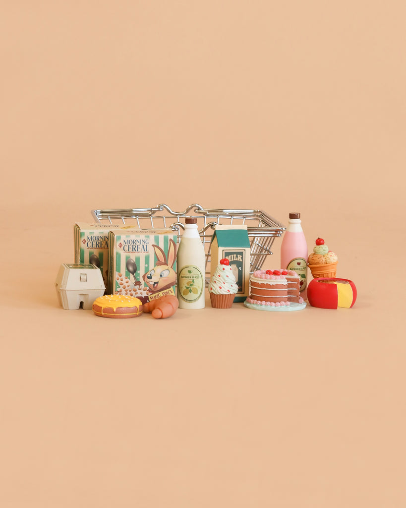 A Konges Sløjd Mini Market Grocery Set featuring a miniature shopping basket surrounded by tiny toy groceries—cereal boxes, milk bottles, eggs, pastries, waffles, a cake, and juice—all displayed against a plain beige background.