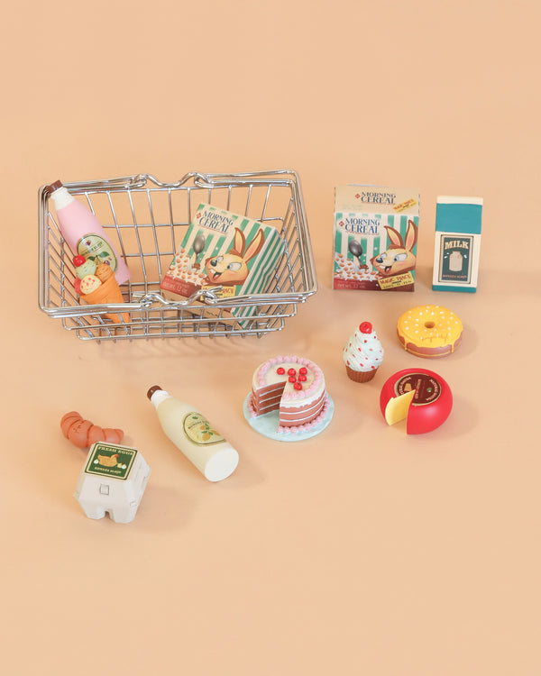 A small wire basket from the Konges Sløjd Mini Market Grocery Set is filled with play food—including toy groceries such as milk, eggs, cookies, cake, cupcake, donut, cheese, juice, and two bunny-themed snack boxes—all arranged on a peach background.