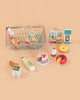 A small wire basket from the Konges Sløjd Mini Market Grocery Set is filled with play food—including toy groceries such as milk, eggs, cookies, cake, cupcake, donut, cheese, juice, and two bunny-themed snack boxes—all arranged on a peach background.