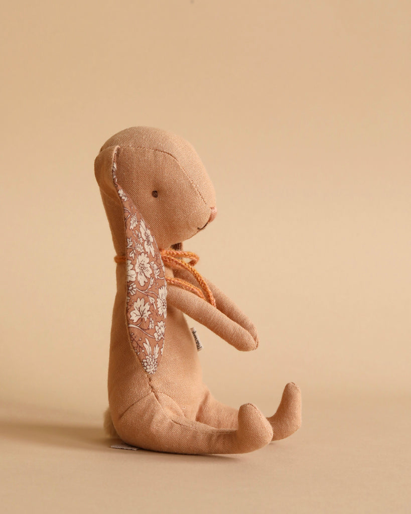 Side view of a Maileg Bunny - Powder sitting upright against a beige background. The toy is made of recycled polyester with a floral pattern on its inner ears and an orange ribbon around its neck. Its limbs are relaxed, and it has a minimalist facial expression.