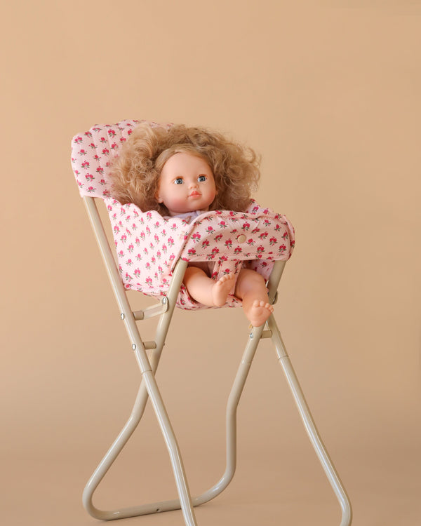 The Konges Sloejd High Chair - Rosie Rose features a curly-haired doll in a pink floral outfit seated in a foldable, detachable high chair, with bare feet dangling and a neutral expression, set against a beige background.