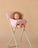 The Konges Sloejd High Chair - Rosie Rose features a curly-haired doll in a pink floral outfit seated in a foldable, detachable high chair, with bare feet dangling and a neutral expression, set against a beige background.