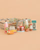 A Konges Sløjd Mini Market Grocery Set small shopping basket filled with toy milk, eggs, bottles, cereal boxes, a cake, a cupcake, an ice cream cone, and cookies is displayed on a beige background.