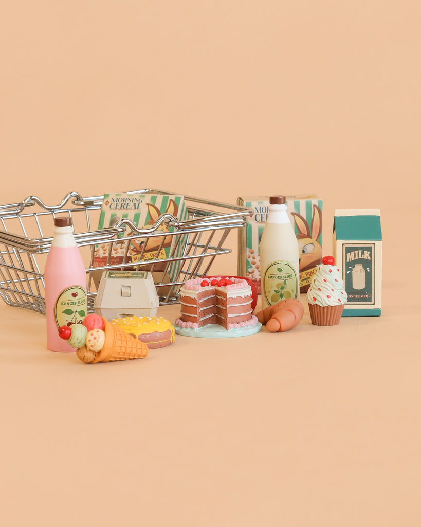 A Konges Sløjd Mini Market Grocery Set small shopping basket filled with toy milk, eggs, bottles, cereal boxes, a cake, a cupcake, an ice cream cone, and cookies is displayed on a beige background.