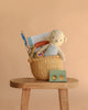 The Easter Basket Gift Set sits on a wooden stool, filled with a Cuddle + Kind Benjamin the Lamb, books, and a toy carrot. In front is a small green box with yellow polka dots for Easter treats, all set against a beige background.