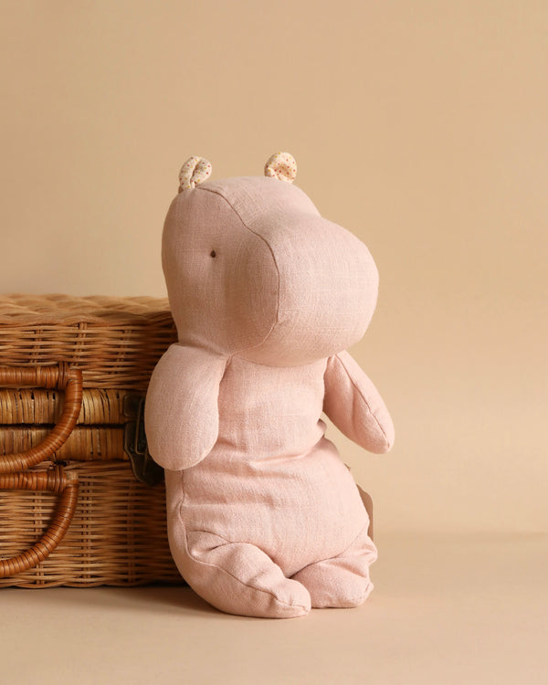 The Maileg Medium Hippo Stuffed Animal in Light Rose, crafted from recycled polyester, leans gently against a woven wicker basket with a beige backdrop. Its simplistic design features small ears and stitched facial details, making it one of the most charming Safari friends.