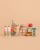 A stuffed mouse in a red striped shirt holds a carrot and sits beside a cake on a stool, surrounded by toy milk bottles, a cereal box, and various play food items from the Konges Sløjd Mini Market Grocery Set, all displayed on a beige background.