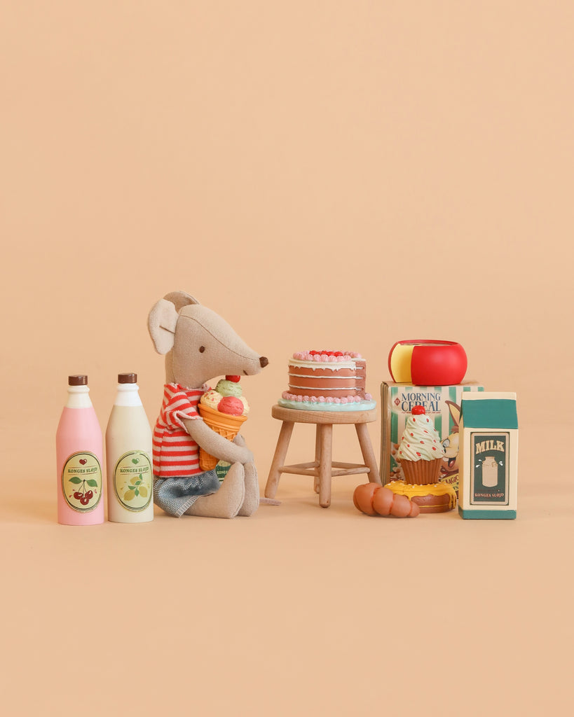 A stuffed mouse in a red striped shirt holds a carrot and sits beside a cake on a stool, surrounded by toy milk bottles, a cereal box, and various play food items from the Konges Sløjd Mini Market Grocery Set, all displayed on a beige background.