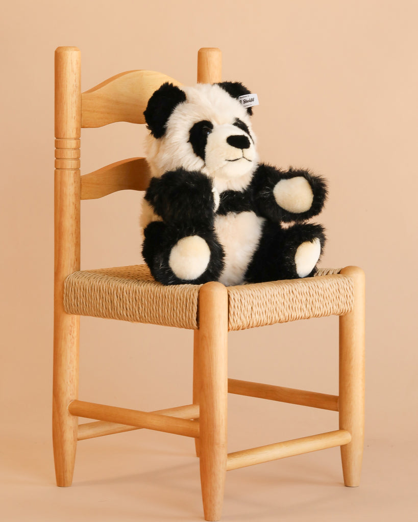 The Steiff Manschli Panda, 9 inches, in black and white plush, sits on a light wooden chair with a woven seat against a plain beige background.