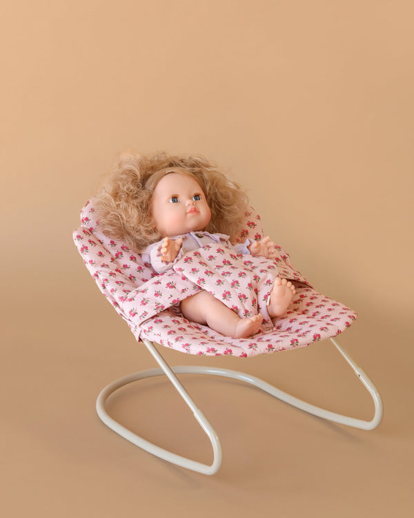The Konges Sloejd Doll Bouncer - Rosie Rose features a curly-haired doll in a floral dress seated in a matching floral-patterned bouncer with a white metal frame, set against a beige background.