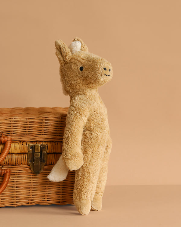 The Senger Naturwelt Stuffed Animal - Beige Horse, crafted from GOTS-certified plush with a white tail and snout, leans against a wicker basket with a brass latch on a plain beige background.