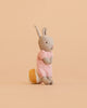 The Maileg Rabbit With Accessories, Micro - Sand - Rose stands upright on a beige background, featuring soft gray fur and a pink knitted dress. Displayed next to a yellow and white round accessory, it’s the perfect addition to a charming rabbit gift set or for inspiring imaginative play as an adorable storytelling toy.