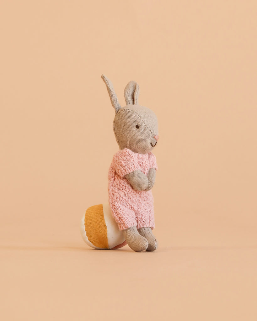 The Maileg Rabbit With Accessories, Micro - Sand - Rose stands upright on a beige background, featuring soft gray fur and a pink knitted dress. Displayed next to a yellow and white round accessory, it’s the perfect addition to a charming rabbit gift set or for inspiring imaginative play as an adorable storytelling toy.