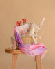 The Easter Basket Gift Set sits on a wooden stool, featuring a plush Maileg Unicorn, Ballet Suit doll, pink flowers, children’s book, rolled poster, and a pink-purple scarf against a beige background.