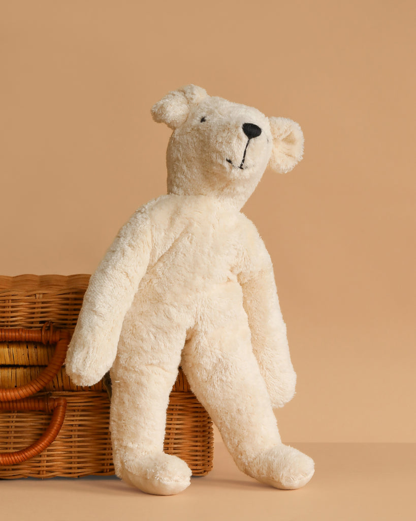 The Senger Naturwelt Stuffed Animal - White Bear, with floppy arms and legs, leans against a wicker basket on a beige background. This charming and eco-friendly plush toy is perfect for sustainable cuddles.