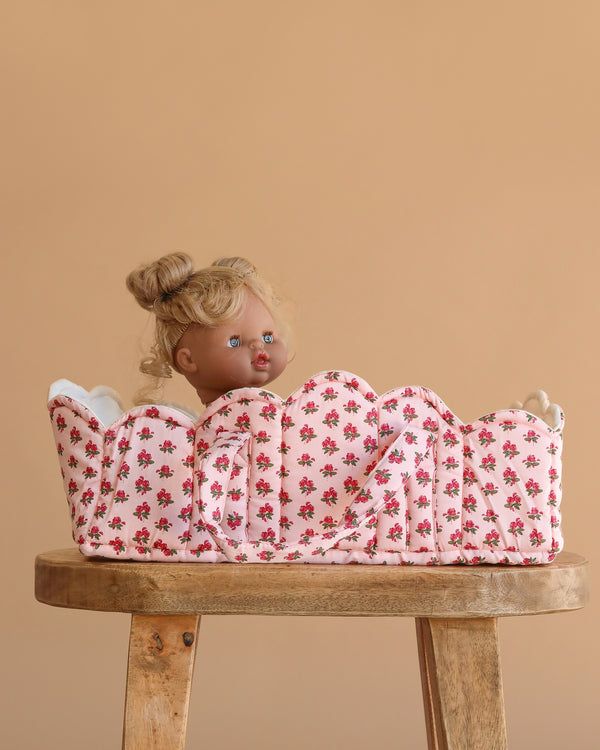 The Konges Sloejd Doll Lift - Rosie Rose, an OEKO-TEX certified floral cotton carrier, holds a blonde blue-eyed doll upright on a wooden stool against a beige background.
