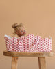 The Konges Sloejd Doll Lift - Rosie Rose, an OEKO-TEX certified floral cotton carrier, holds a blonde blue-eyed doll upright on a wooden stool against a beige background.