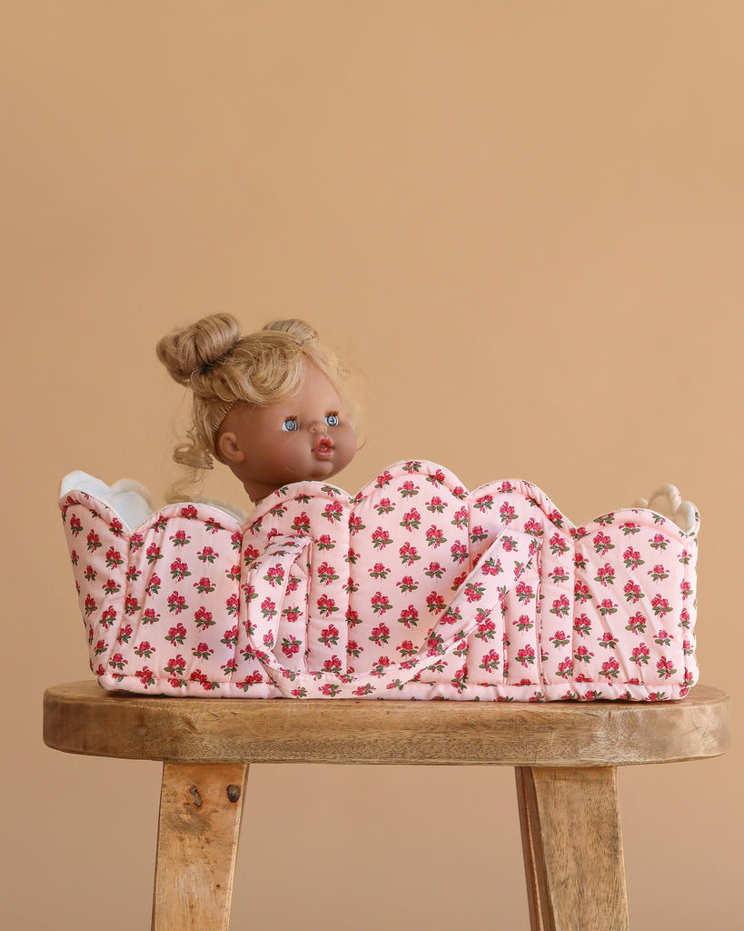 The Konges Sloejd Doll Lift - Rosie Rose, an OEKO-TEX certified floral cotton carrier, holds a blonde blue-eyed doll upright on a wooden stool against a beige background.