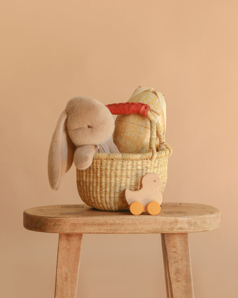 The Easter Basket Gift Set, featuring a Maileg Plush Bunny, patterned cushion, and wooden toy in a woven basket on a stool, is displayed with a Rolling Duck on wheels against a solid beige background.