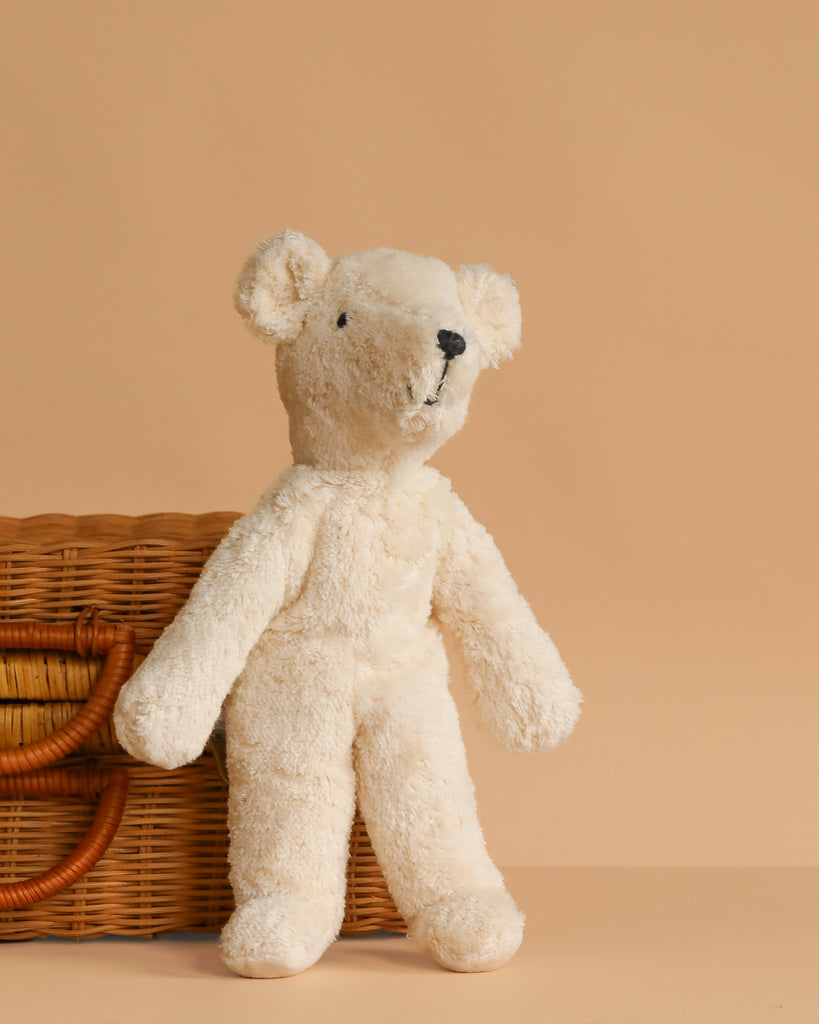 A soft, cream-colored Senger Naturwelt Stuffed Animal - White Bear stands upright by a wicker basket with curved handles on a beige background—a perfect sustainable addition to any nursery.