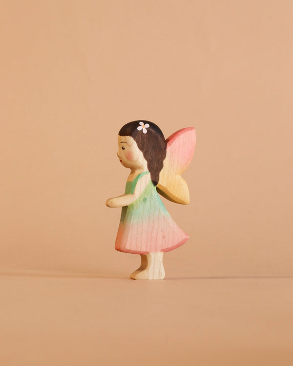 A Handmade Holzwald Maya Fairy, featuring brown hair adorned with a flower, a pastel green and pink dress, and yellow-pink wings, stands against a beige background. This enchanting piece comes with a postcard and is part of our collection of handmade wooden figures—perfect for imaginative play or whimsical decoration.