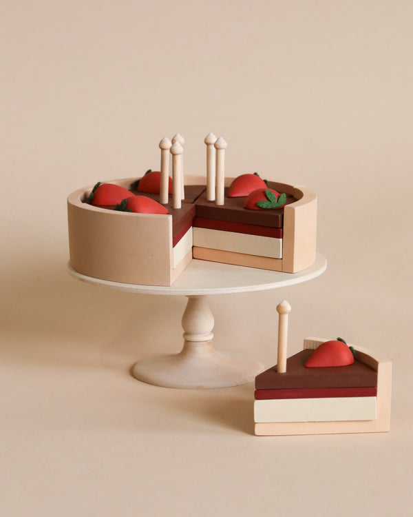 A wooden cake stand displaying a Handmade Chocolate Layer Cake On A Stand divided into slices, each topped with a red apple and white candles, on a beige background. The toy is crafted from non-toxic paint.