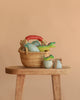 The Easter Basket Gift Set, filled with colorful dinosaur toys, sits on a wooden stool alongside two dinosaur-themed jars and a Handmade Wooden T-Rex, all against a light beige background.