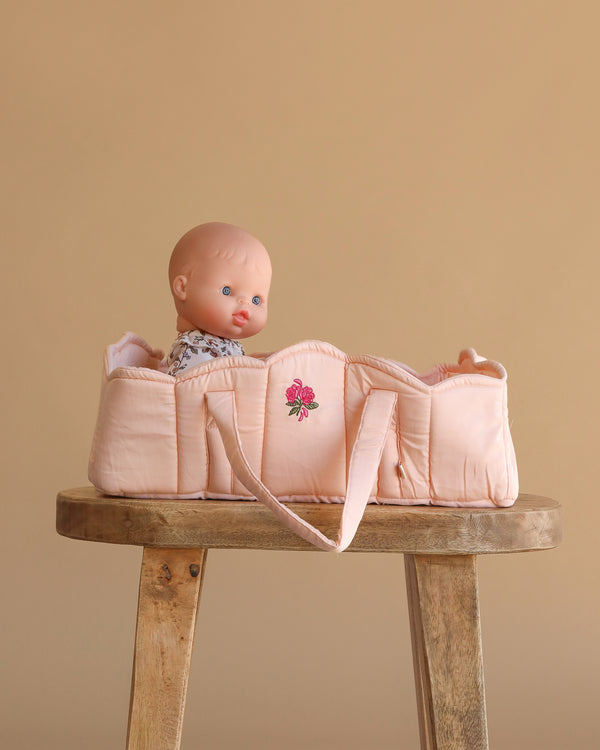 A baby doll sits upright in the Konges Sloejd Doll Lift - Cameo Rose, a soft, pink OEKO-TEX certified cotton carrier with embroidered flower details, placed on a wooden stool against a plain beige background.
