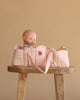 A baby doll sits upright in the Konges Sloejd Doll Lift - Cameo Rose, a soft, pink OEKO-TEX certified cotton carrier with embroidered flower details, placed on a wooden stool against a plain beige background.