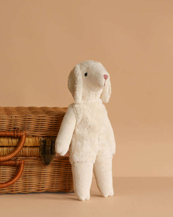 The Senger Naturwelt Stuffed Animal - Small Sheep, GOTS-certified and white, stands upright beside a latched wicker basket against a beige background.