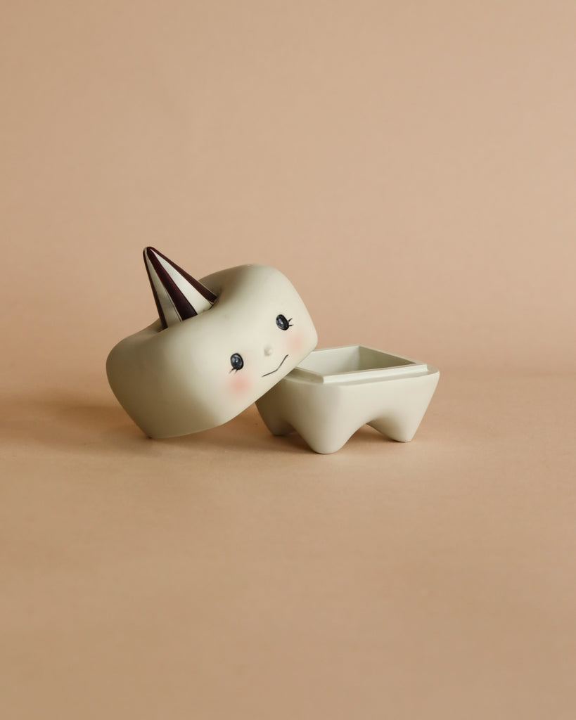 A small, beige ceramic container with a cute face and blush cheeks, shaped like a tooth. Perfect for holding a child's first teeth, its lid is slightly ajar, revealing a square compartment inside. The Konges Sløjd Stone Tooth Box sports a brown and white striped conical cap on top. The background is a soft beige.