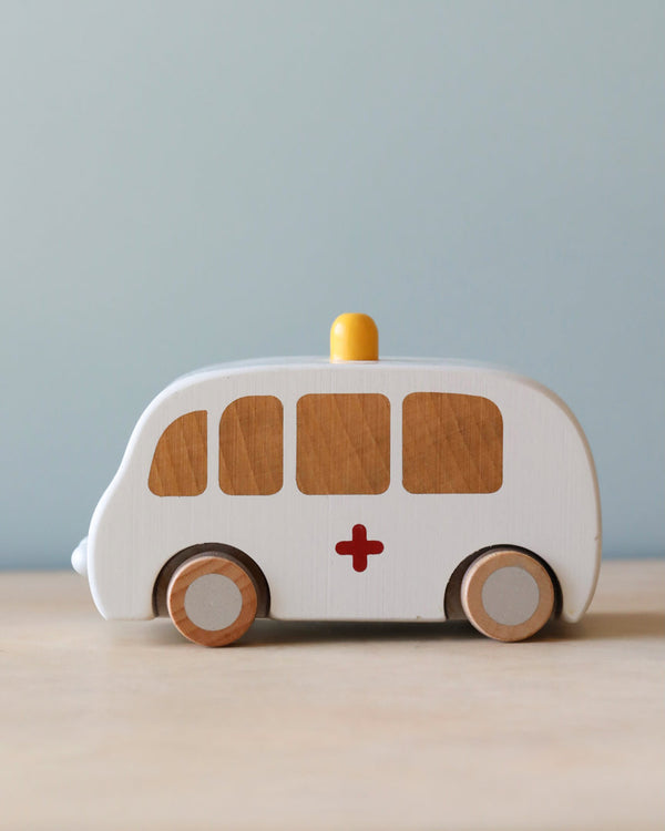 The Maileg Wooden Ambulance has a solid wood white body, brown windows, and a red cross. It rolls on wooden wheels with a yellow siren on top, set against a light blue background.