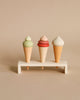 Three handmade ice cream cone toys with green, pink, and white swirls on top, displayed on a tiny wooden shelf against a neutral background.
