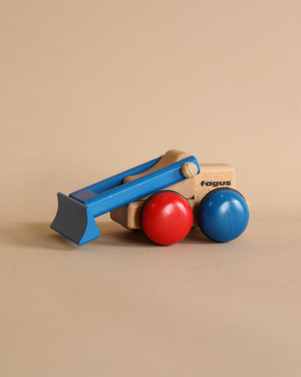 A wooden Fagus Wooden Bulldozer - Mini Series handcrafted in blue, red, and natural wood colors against a plain beige background. The bulldozer features a blue hatchback design with rounded red and blue wheels.