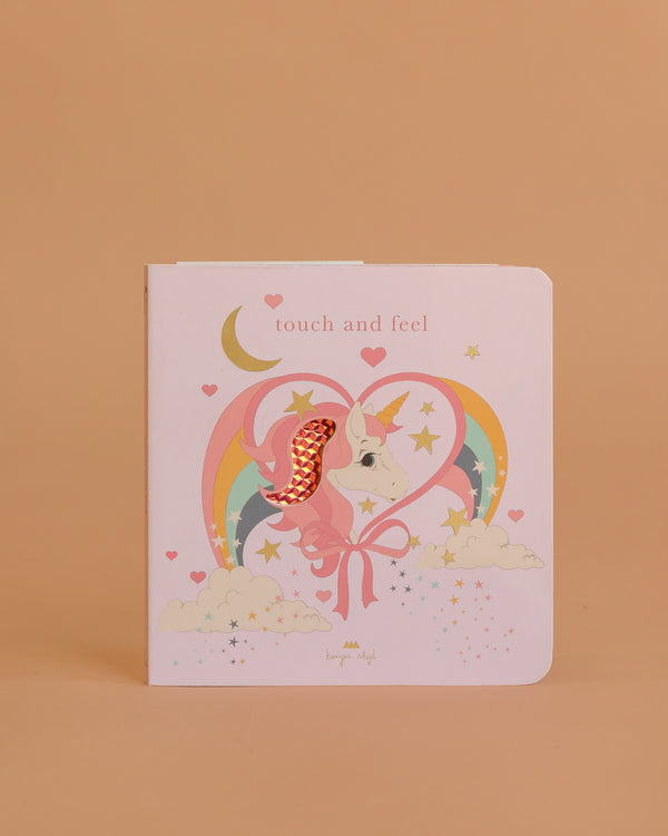 The Konges Sloejd Touch and Feel Book - Unicorn features a pastel pink cover with a unicorn, sparkly mane, rainbow, stars, clouds, and hearts on a peach background. This sensory cardboard book is perfect for young children to explore.