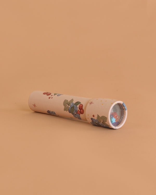 The Konges Sloejd Kaleidoscope - Cremona, featuring a floral pattern with berries, leaves, and colorful pearls, rests on a peach-colored surface with its viewing end angled slightly to the right.