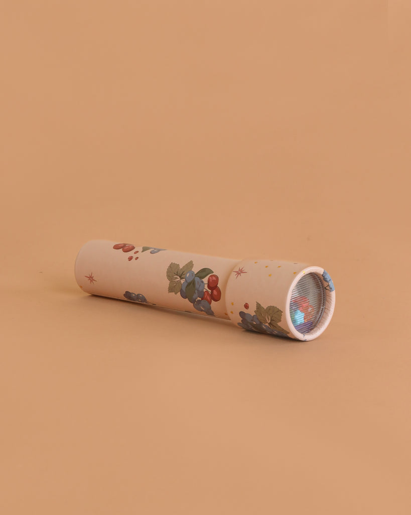 The Konges Sloejd Kaleidoscope - Cremona, featuring a floral pattern with berries, leaves, and colorful pearls, rests on a peach-colored surface with its viewing end angled slightly to the right.