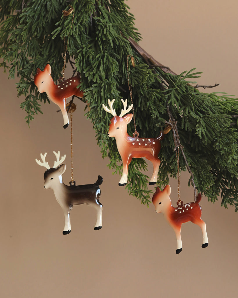 Four Maileg Reindeer Family metal ornaments with antlers hang gracefully from a lush green pine branch. In various shades of brown and white, they add a festive touch to the scene. Perfect as a family gift, their charm is enhanced against a neutral beige background.