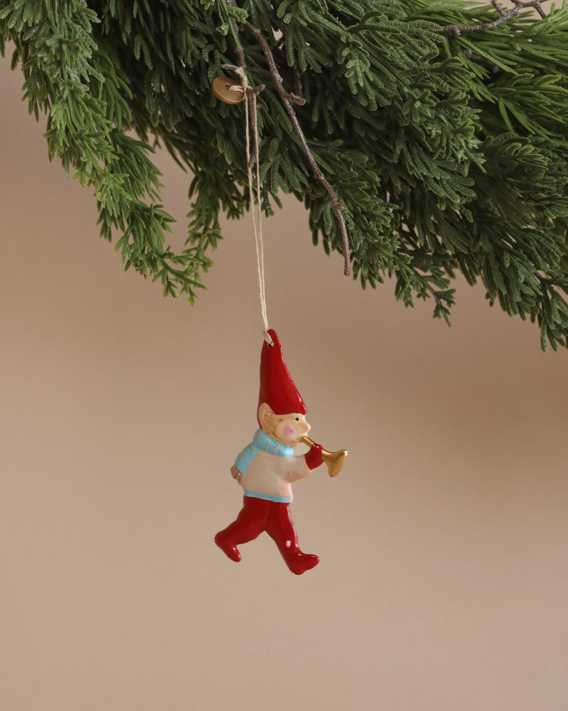 A whimsical Maileg Metal Ornament, Pixy Trumpet, featuring a gnome in a red hat and pants holding a small horn, hangs from a green pine branch. Handpainted details accentuate its charm against the soft beige background.