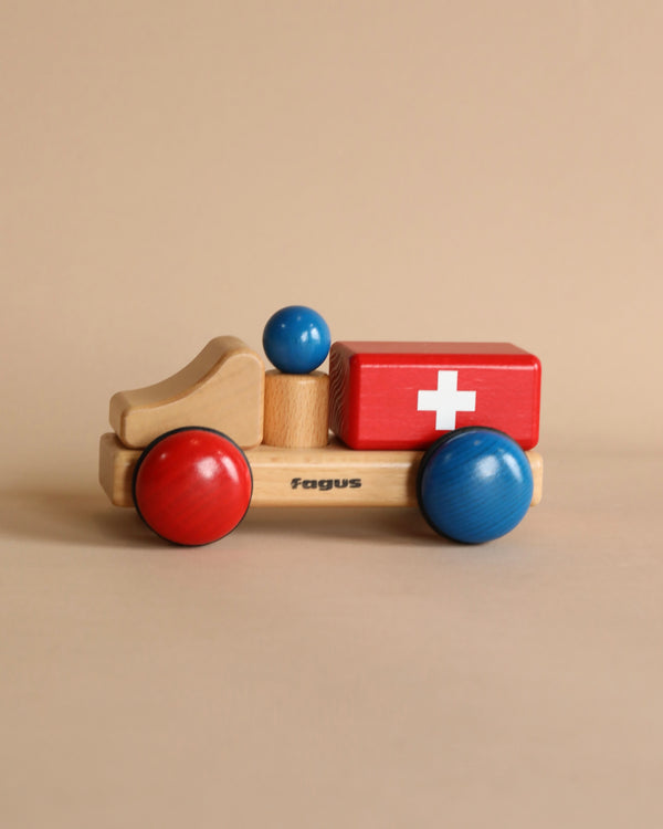 A Fagus Wooden Ambulance - Mini Series with blue wheels, a red body featuring a white cross, and a natural wood driver's cabin on a beige background. The word "Fagus" is visible on the ambulance.