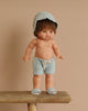 The Minikane Standing Doll (14") - Julian With Clothing features an anatomically correct baby doll with short brown hair, dressed in a light blue hood, matching shorts, and slippers, posed on a wooden bench against a beige background.