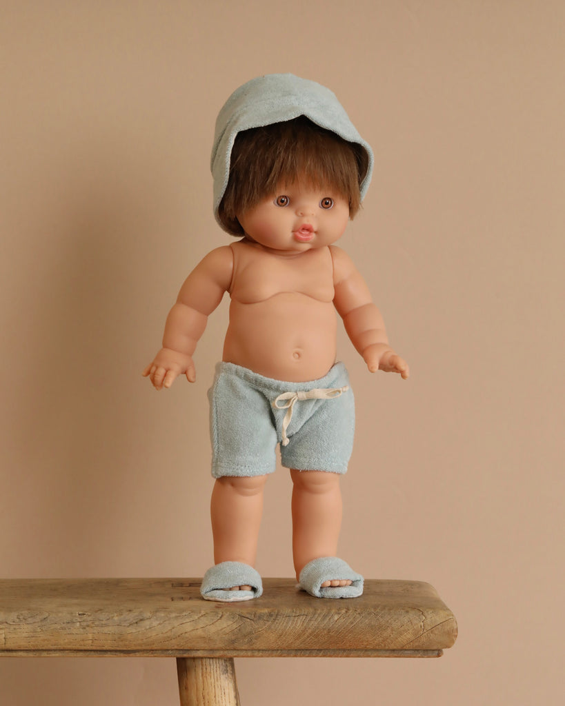 The Minikane Standing Doll (14") - Julian With Clothing features an anatomically correct baby doll with short brown hair, dressed in a light blue hood, matching shorts, and slippers, posed on a wooden bench against a beige background.