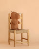 A Maileg Rhino, Medium - Chocolate, crafted from soft fabric in a warm brown hue, sits upright on a wooden chair with a woven seat against a plain beige background—a charming addition to your collection of Safari friends.