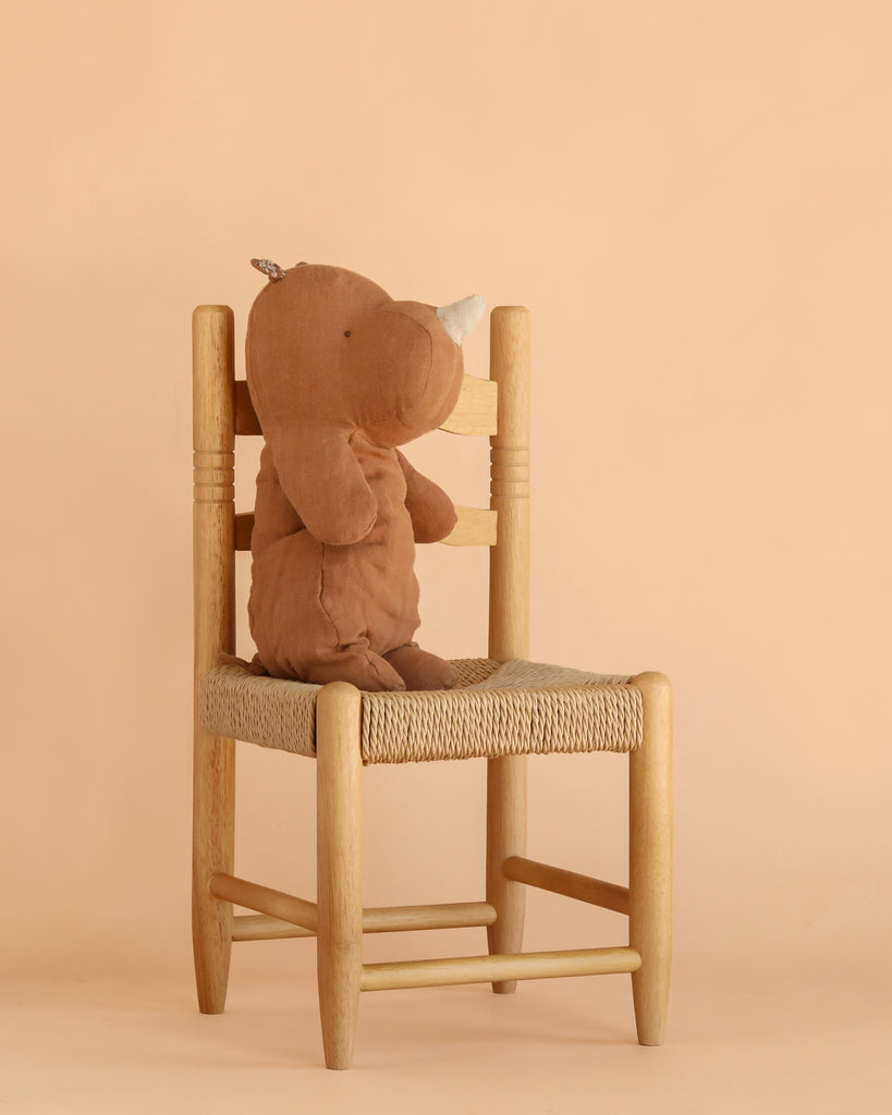 A Maileg Rhino, Medium - Chocolate, crafted from soft fabric in a warm brown hue, sits upright on a wooden chair with a woven seat against a plain beige background—a charming addition to your collection of Safari friends.