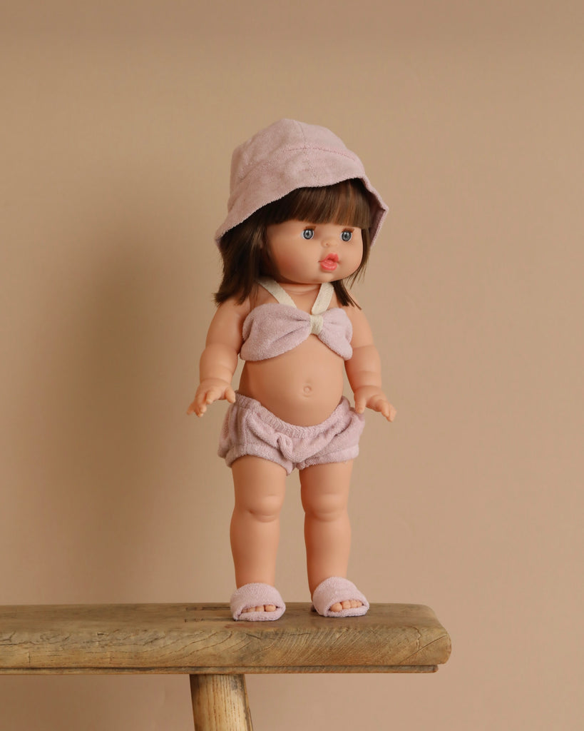 The Minikane Standing Doll (14") - Chléa With Clothing features brown hair with bangs and comes dressed in a pale pink hat, top, shorts, and slippers. This anatomically correct doll stands on a wooden bench against a beige background.