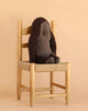 A Maileg Gorilla, Medium from the Safari Friends collection sits on a light wooden chair with a woven seat, against a beige background. Crafted from recycled polyester, this plush gorilla toy is an eco-friendly stuffed animal option.