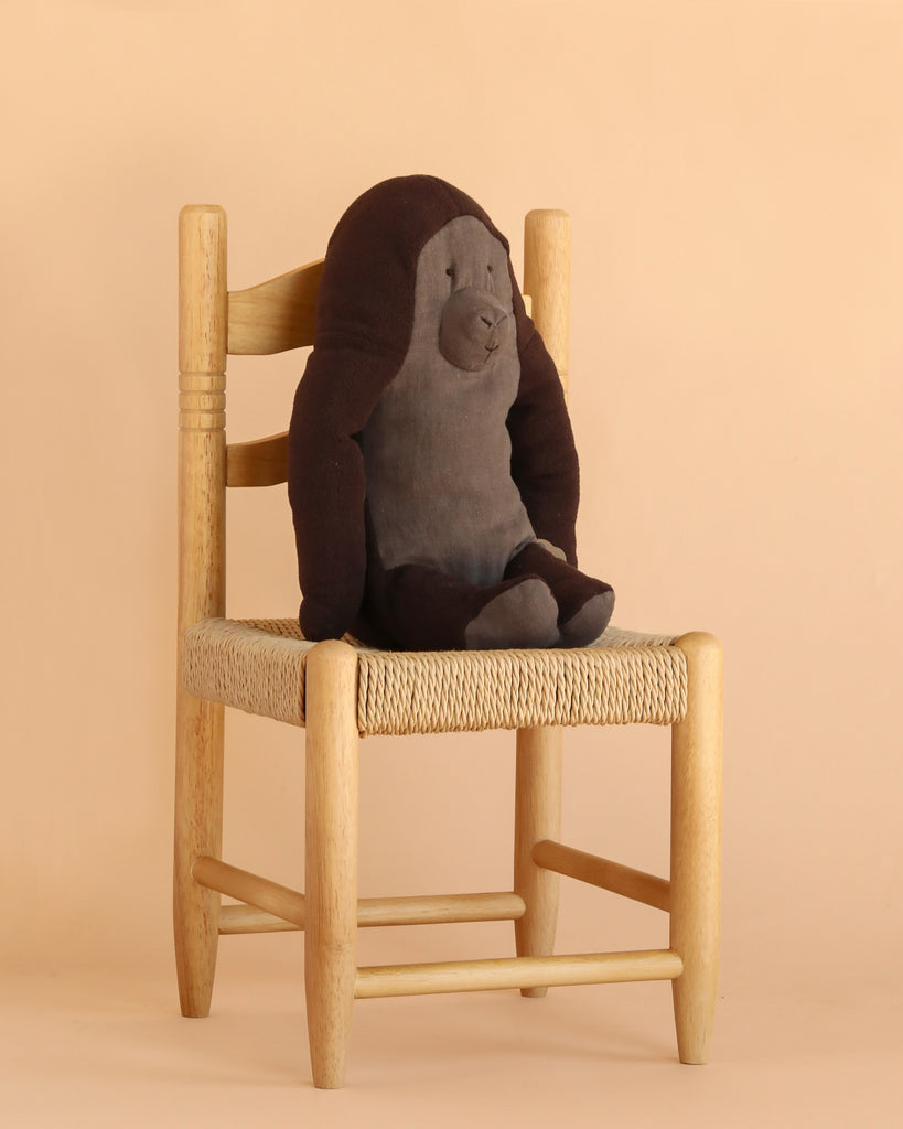 A Maileg Gorilla, Medium from the Safari Friends collection sits on a light wooden chair with a woven seat, against a beige background. Crafted from recycled polyester, this plush gorilla toy is an eco-friendly stuffed animal option.